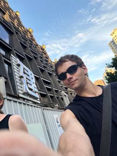 This image is a bright, upward-angled selfie taken by a man in an urban setting. The man, with dark hair and wearing a black T-shirt and black sunglasses, occupies the upper part of the frame, looking towards the camera with a relaxed expression. A black strap, possibly from a bag, is visible across his shoulder. Partially in the bottom right corner, a glimpse of another person's head and shoulder can be seen.

The background prominently features a modern building with a distinctive architectural facade, characterized by numerous angular, layered, grey structures, possibly for sun shading. Small green plants are visible on some of the building's ledges. A large, stylized metallic sign, appearing to be three glossy silver characters or a logo, is affixed to this building, though the exact text is not clearly legible but suggests "MIA" or similar stylized Chinese characters.

Above the buildings, a clear blue sky with scattered wispy white clouds indicates a sunny day, likely midday or afternoon. In the distance, other light-colored buildings are visible. The setting depicts a vibrant outdoor urban environment in Putuo District, China. A corrugated metal fence with an illegible white sign is partially visible on the right side of the image.