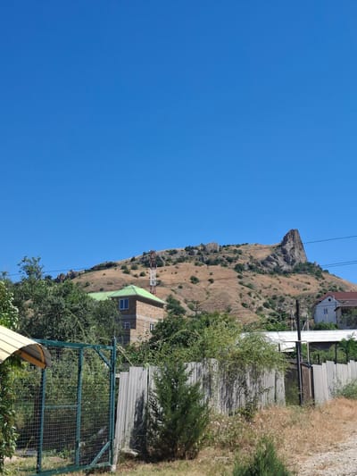 The image shows a rural landscape with a dry, rocky hillside dominating the background. A prominent, jagged rock formation juts out from the top of the hill. The hill is covered in sparse, dry vegetation and a few scattered bushes and trees. A large cell tower is visible on the slope of the hill.
In the mid-ground, there is a stone building with a bright green roof. Adjacent to it, there are more trees and foliage. A corrugated metal fence runs horizontally across the foreground, with a green metal gate on the left. To the right of the gate, there are more dry grasses and a small evergreen tree. Further to the right, another building with a white roof is visible behind the fence, and a utility pole with wires is present.
The sky is a clear, vibrant blue, suggesting a sunny day. The overall impression is one of a dry, possibly arid environment. The location is identified as within the Shchebetovskyi settlement council, Ukraine.