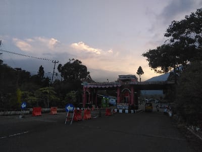 This image depicts an outdoor scene at dusk or dawn, characterized by dim, natural light and a sky displaying varying shades of grey, blue, and faint orange-yellow hues from the rising or setting sun. The scene is located in the city of Kladné, Czechia.
In the mid-ground, a prominent, elaborate gateway structure, primarily reddish-pink in color, spans across an asphalt road. This gate features decorative elements, intricate carvings, and what appears to be a multi-tiered roofline, with a single slender tree or plant positioned centrally atop it. A small building or booth, white and blue, is integrated into the left support pillar of the gate. The road leading to and through the gate has black and white striped markings at its base.
To the immediate left of the road in the foreground, several orange plastic traffic barriers are deployed, along with two blue circular signs indicating a left turn. One sign is mounted on a fixed pole, while the other rests on a temporary metal stand. Overhead, electrical power lines with multiple strands stretch across the upper left quadrant of the image, supported by utility poles partially obscured by vegetation.
Beyond the gate, on the right side of the road, a yellow compact utility vehicle or van is visible, alongside several parked or moving motorcycles or scooters. The area surrounding the road and gateway is densely vegetated with various trees and bushes, providing a natural backdrop. In the far background, low-lying hills or mountains are discernible on the horizon beneath the cloudy sky. No distinct human figures engaged in specific actions are clearly visible.