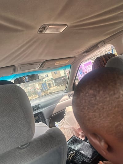 The image depicts an interior view from the rear seat of a vehicle, looking forward through the windshield onto a street in Eneka, Nigeria.
Inside the vehicle, a light-colored fabric headliner with a dome light fixture is visible at the top. Below this are two sun visors, the passenger-side visor partially deployed with a warning label. A rearview mirror centrally reflects parts of the vehicle's interior and a person. The dark-toned dashboard and a stereo unit are visible in the foreground. Gray fabric car seats occupy the middle ground; the top of the front passenger seat's headrest is visible.
Occupants include: an individual in the front passenger seat, dressed in light-colored clothing; a person with dark, short hair partially obscuring the lower right foreground, holding a black, textured purse with a gold chain strap, and appearing to look downwards; and a person with braided brown hair visible behind the front passenger. The driver is not directly visible but reflected in the rearview mirror.
Outside the vehicle, the street scene in Eneka reveals multiple structures under bright daylight. A two-story building with white walls and a darker upper section is prominent on the left. Further right, roadside activity includes a person with a blue container, possibly on a cart, and various other objects suggesting vendor stalls. A dark-colored vehicle is partially visible further down the street. A large, colorful advertising banner or billboard is visible on the right side of the road, featuring graphics and text.