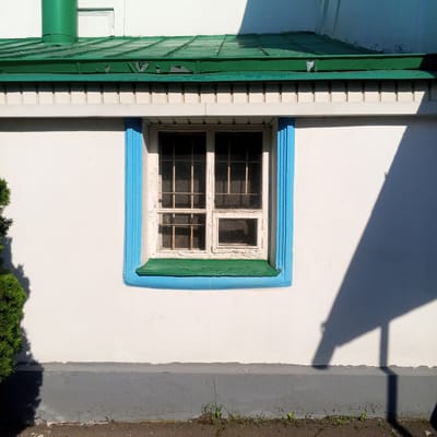 A close-up, eye-level, exterior shot focuses on a white, plastered wall of a building in Zhilino-1, Russia. The wall is interrupted by a window with a blue frame and a green sill. The window has six panes, each with a metal grate. The paint on the window frame is peeling. Above the window, there is a white trim with vertical grooves, and above that, a green, corrugated metal roof with peeling paint. A green pipe protrudes from the roof to the left. The base of the wall is painted gray and shows signs of wear. To the left of the frame, a portion of a dark green evergreen shrub is visible, casting a shadow. To the right, a large, dark shadow, likely from an overhanging structure, is cast across the wall. The ground in front of the wall is a rough, dark surface, possibly asphalt or gravel, with some sparse green weeds growing near the base of the wall. The lighting suggests it is daytime, with strong sunlight creating distinct shadows. No people or animals are present in the image.