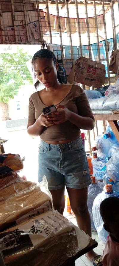 In Kwachiri, Nigeria, a young woman with braided hair stands in what appears to be an outdoor market stall. She is wearing a brown, off-the-shoulder top and denim shorts, holding a smartphone in her hands and looking down at the screen. The stall is stocked with various goods, including loaves of bread wrapped in plastic, plastic water bottles, and what look like bags of flour or other dry goods. Behind the woman, posters or advertisements are tacked to a grid-like structure. The environment suggests a busy, possibly warm, day. The overall impression is a snapshot of daily life and commerce in a Nigerian urban setting.