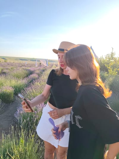 Two young women are in a lavender field in Nechaiane, Ukraine, likely at sunset. The woman in the foreground is wearing a black t-shirt with a faded grey graphic design and a watch on her left wrist. She is holding a purple hairbrush in her right hand. The woman behind her is wearing a black t-shirt and a straw hat. She is holding a smartphone in her left hand, appearing to take a selfie with the younger woman. The setting is a vibrant lavender field with rows of purple flowers stretching into the distance. In the background, there are some white tent-like structures, possibly set up for an event or activity. The sun is low in the sky, casting a warm, golden light on the scene, creating lens flare and highlighting the hair of the woman in the foreground. The sky is a clear blue. The overall mood appears relaxed and enjoyable, with the women engaged in a moment of leisure and likely capturing memories.