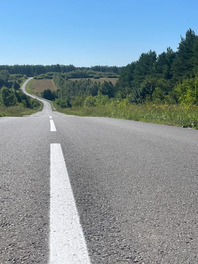 The image shows a winding asphalt road with a white dashed line marking the center. The road is bordered by green grass and trees on either side. In the background, there are rolling hills covered with more trees. The sky is clear and blue. The scene appears to be a rural or suburban area in Maloe Murashkino, Russia. There are no people or vehicles visible in the image.