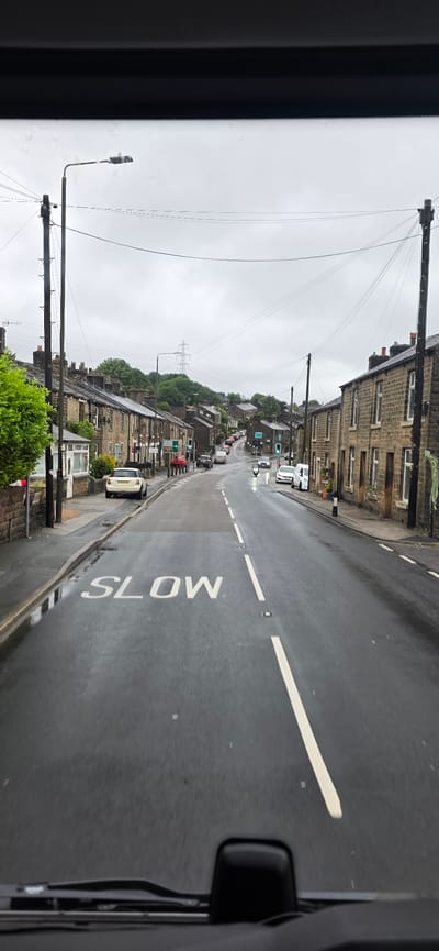 The image depicts a street scene in High Peak, United Kingdom, on an overcast day. The road surface is wet, indicating recent rain, and features a "SLOW" marking painted in white lettering on the asphalt. To the left of the road, a white car is parked. The street is lined with terraced houses constructed of stone, with multiple windows visible. A street lamp stands on the left side of the road, and power lines stretch across the sky. Further down the street, other vehicles are visible, including cars, a van, and a motorcycle. Traffic lights are also present, showing a green signal. Trees and vegetation are interspersed with the houses, and a tall pylon is visible on a distant hillside. The image appears to have been taken from inside a vehicle, as the dashboard and windshield wipers are visible at the bottom.