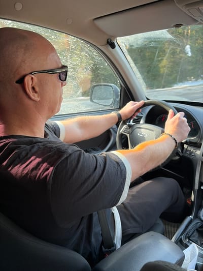 An overhead-view forensic image captures the interior of a vehicle in motion, specifically the driver's position, in Zabegalovo, Russia. The driver is a bald male, viewed from behind his right shoulder, wearing dark sunglasses, a dark short-sleeved t-shirt with light cuffs, and dark trousers. His left wrist bears a dark watch, and his right wrist has a dark beaded bracelet. He is seated in a dark upholstered seat with his seatbelt fastened, and both hands are positioned on the black steering wheel, which features a silver emblem, likely a Chery logo. The dashboard and a portion of the instrument cluster with red needles are visible through the steering wheel. An automatic gear shifter is visible to the driver's right. The windshield and passenger side window are visible, appearing somewhat splattered with debris, possibly road grime or melted snow. Through the windows, a tree-lined, possibly forested, exterior environment is visible, with patches of snow on the ground, indicating a cold or wintery setting. Sunlight illuminates parts of the scene, casting shadows on the driver's shirt and arms. A small black device, possibly a microphone, is attached to the vehicle's interior near the top right of the windshield.