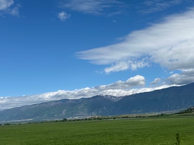 A landscape shot, oriented vertically, depicts a rural scene dominated by the sky and distant mountains. The sky, a deep blue, features elongated white clouds stretching across the lower half of the image. Above the clouds, a mountain range stretches horizontally. At the base of the mountains, a narrow strip of dense trees and a town appear before a field of green grass. The scene is Marino Pole, Bulgaria.