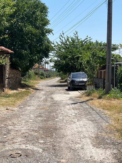 A grey Skoda Karoq is parked on a dirt road in Akandzhievo, Bulgaria. The car is facing forward and is partially obscured by trees. The road is lined with stone walls and houses, and there are power lines overhead. The sky is clear and blue. The car's license plate reads "CB 2355 AE".