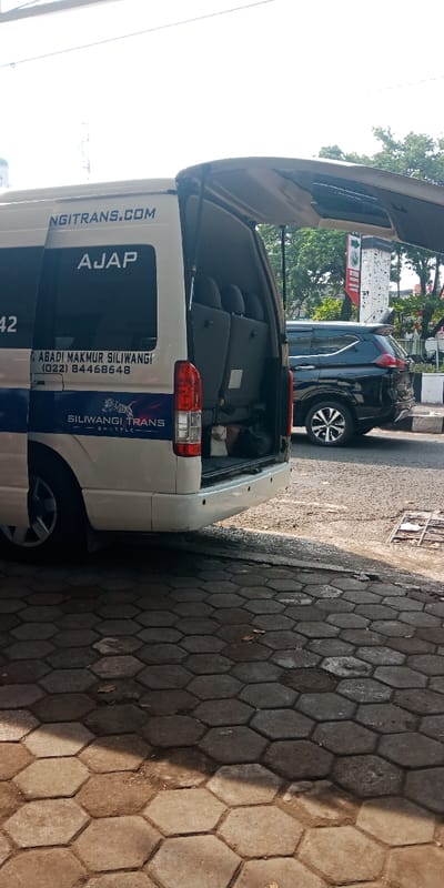 The scene depicts a parked white passenger shuttle van, identified by "SILIWANGI TRANS SHUTTLE" and "NGITRANS.COM" branding, located in Bojongherang, Indonesia. The van's rear hatch is fully open, revealing multiple rows of unoccupied grey passenger seats and a black bag in the cargo area. A phone number, (022) 84468648, and "ABADI MAKMUR SILIWANGI" are visible on its side. The ground in the foreground is paved with hexagonal concrete blocks, showing variations in shade and minor staining. Directly behind the van, a dark-colored SUV is parked on an asphalt road, partially obscured. In the background, green foliage from trees, a utility pole, and a red and white street sign with text and directional arrows are visible. The setting is outdoors, under natural daylight, casting shadows on the paved ground. No individuals are visible in the immediate vicinity of the van.