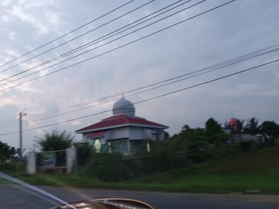The image shows a mosque with a red roof and a silver dome, located in the city of Cotbuket, Indonesia. The mosque is surrounded by greenery, and there are power lines visible in the foreground. The sky is overcast with clouds. The photo appears to be taken from a moving vehicle, as indicated by the blurriness of the foreground and the dynamic perspective. The overall atmosphere is calm and serene.