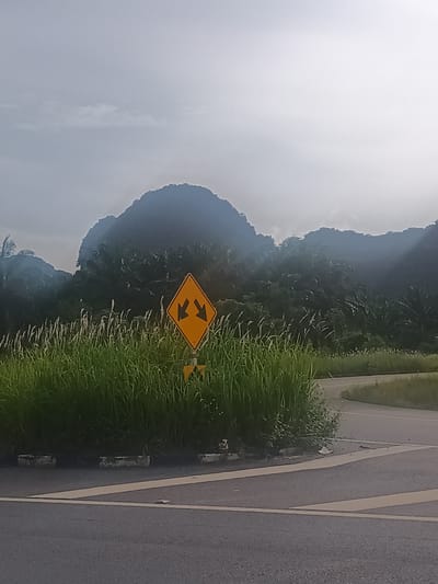 The image shows a road junction in Ban Klang, Thailand. A yellow diamond-shaped road sign with black arrows pointing left and right is positioned in the center of a grassy island separating traffic lanes. The sign is partially obscured by tall grass. Behind the sign, a large, dark, misty mountain or hill dominates the background. The sky is overcast, and the road surface is asphalt with painted white lines. No people are visible. The scene suggests a rural or semi-rural location with mountainous terrain.