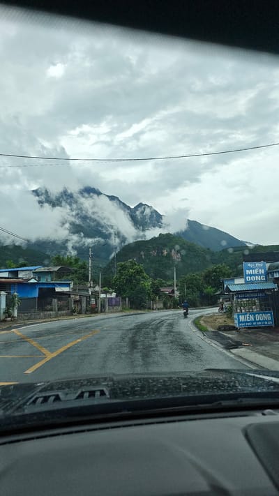 The image, taken from inside a vehicle, shows a street scene in what appears to be the city of Bình Lư, Vietnam, based on the signage visible. The road is wet, likely from rain, and marked with yellow lines. The mountains in the background are shrouded in clouds. Buildings and greenery line the side of the road. A sign on the right side of the road reads "Học Phú MIẾN DONG" with a phone number. A person on a motorbike is visible on the road. The sky is overcast. The dashboard of the car is visible at the bottom of the frame.