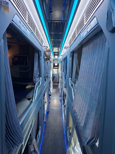 The image shows the interior of a sleeper bus, likely in Tân Phú 2, Vietnam, with narrow sleeping berths arranged on either side of a central aisle. The berths are enclosed with gray curtains, and some have small screens mounted inside. Blue LED lights run along the ceiling and base of the berths, illuminating the corridor. A small, brown, stuffed toy with a smiley face is visible hanging from the side of a berth. The floor is made of wooden planks. The overall impression is one of a compact, modern sleeping arrangement for travel.
