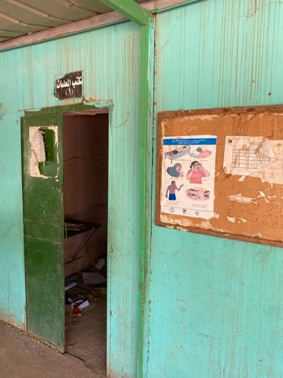 The image shows the exterior of a building in Khartoum, Sudan, located at coordinates 15.552, 32.57. The building is constructed with light blue painted wood panels. The building appears to be in a state of disrepair with peeling paint and rusty metal accents. There is a green door leading to a room with a single desk and chairs. There is a bulletin board on the wall with a poster of a man and woman with medical instructions written in Arabic. The bulletin board also has a grid of boxes filled with numbers.  There is a sign above the doorway which says "غرفة المعاينات." The room is cluttered with various objects including a few sheets of paper scattered on the floor.  The door is slightly ajar, revealing a glimpse of the interior. The image appears to be taken from a distance, and the photographer is positioned to the left of the building. The photo was likely taken during the daytime.