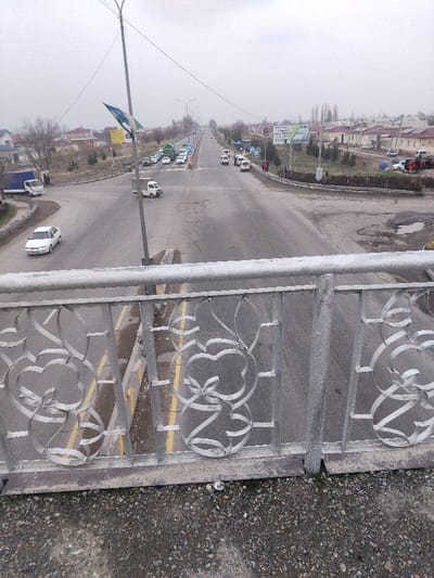 An elevated viewpoint captures an overcast daytime scene of a road network in Tashlak, Uzbekistan. In the immediate foreground, a weathered, light-gray painted metal bridge railing featuring decorative, intertwined scrollwork is visible. Below the railing, a gravel-strewn surface with sparse vegetation and a discarded silver object (possibly a can) is present.
The midground features an asphalt main road extending longitudinally into the distance, marked by visible cracks, patches, and faint double yellow lines. An asphalt secondary road branches off to the left, forming an intersection. Vehicles are distributed across the scene: a white sedan moves on the left branch road, a white utility truck is positioned near the center of the main road, and a cluster of vehicles, including green trucks and several white cars, are stopped at what appears to be a traffic signal further down the main road. Traffic signals on the right side of the main road display red lights.
A tall utility pole with overhead wires stands prominently near the center-left, bearing a blue, white, and green flag, identified as the flag of Uzbekistan. Multiple streetlights line the main road. On the right side, a large billboard with green-themed graphics and text is visible. Several pedestrians are observed on the right shoulder of the main road, near the stopped vehicles and traffic signals.
The background reveals low-rise buildings with light-colored facades and various roof types on both sides of the thoroughfare. Bare trees and sparse foliage suggest a non-summer season. The sky is uniformly overcast, contributing to a muted light quality across the entire scene.