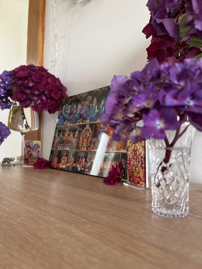 The image depicts a close-up view of a wooden surface with various decorative items. In the foreground, a clear glass vase with a diamond-cut pattern holds purple hydrangeas. To the left, a collection of religious images is displayed on a table, including a framed picture of deities, a small elephant figurine, and a silver wine glass. The background features a white wall with string lights and a wooden post. The overall scene suggests an indoor setting, possibly a home or a shop, decorated with flowers and religious iconography. There are no people visible in the image.