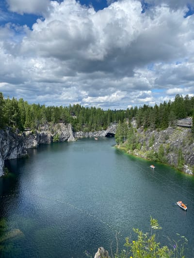 This image captures a scenic view of the Ruskeala Mountain Park in Russia. The park features a picturesque turquoise lake surrounded by steep, rocky cliffs and dense green coniferous forests. The water is calm and reflects the dramatic, cloudy sky above, which is a mix of bright white and dark grey cumulus clouds. Several small white boats with orange-vested occupants are visible on the lake, suggesting recreational activities. One boat is in the foreground, with two people visible, and another is further out in the distance. A line of yellow buoys stretches across a section of the water. Wooden walkways and observation decks are built along the cliff edges, providing vantage points for visitors. The overall scene depicts a serene natural landscape enhanced by human elements of tourism and leisure.