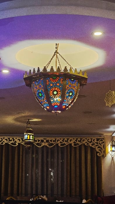 A colorful, ornate chandelier hangs from a purple ceiling with inset lighting. The lamp features intricate patterns of various colors. An octagonal, laser-cut design is visible above the chandelier. Adjacent to it are wooden blinds with decorative trim that frame the side. Another similar lantern hangs near the blinds, contributing to the decorative ambiance.