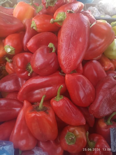A close-up, overhead shot shows a large pile of bright red bell peppers, some with orange hues, at an outdoor market in Razzakov, Kyrgyzstan. The peppers are of various sizes and shapes, with some appearing smooth and glossy, while others have a slightly wrinkled texture. Several green stems are visible, indicating freshness. The lighting is natural and bright, suggesting it is daytime. In the background, a glimpse of white packaging and other produce can be seen, hinting at a vibrant market environment. The image is dated 25.06.2026 at 13:08.
