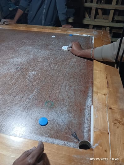 The image depicts a carrom game in progress at 18:49 on February 12, 2025, within the city of Maliat, Bangladesh. The central subject is a wooden carrom board with a reddish-brown playing surface heavily dusted with white carrom powder, showing wear and tear, including cracks in the wooden frame. One circular pocket is visible in the lower right corner. A hand with tan skin, wearing a silver-colored link bracelet, is positioned on the right side of the board, holding a white circular carrom striker, poised to make a shot. The forearm is covered by a light brown sleeve. Two white carrom men and two blue carrom men are scattered across the playing surface. A second hand with a darker skin tone is visible on the lower left wooden frame. In the background, partially obscured, is a third individual, seen from the waist up, wearing a dark blue or gray long-sleeved garment, with their hands resting on the far wooden edge of the carrom board. The background environment is dimly lit, revealing wooden structures or shelving, indicative of an indoor setting.