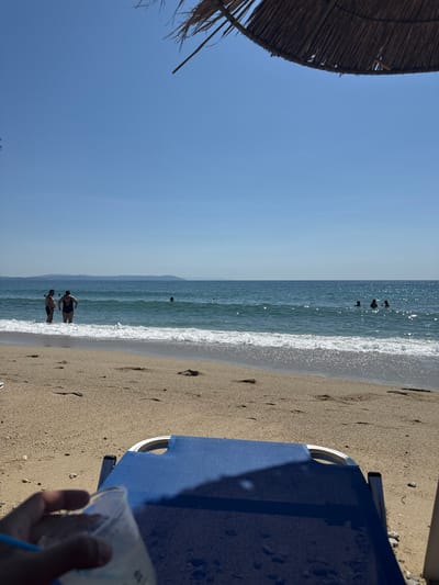 A sunny day at Asprovalta Beach, Greece, is captured from the perspective of someone relaxing on a blue beach chair. In the foreground, a hand holds a clear plastic cup with a blue straw, suggesting a beverage. The sandy beach stretches towards the vibrant turquoise waters of the Aegean Sea, where gentle waves break on the shore. A thatched umbrella provides shade in the upper right corner of the frame.
Further out in the sea, several individuals are visible. Two people stand near the shoreline, with the male wearing dark shorts and the female in a black swimsuit. Other swimmers are scattered throughout the water. In the distance, a hazy mountain range outlines the horizon. The sky is a clear, bright blue, indicating excellent weather conditions for a beach outing.