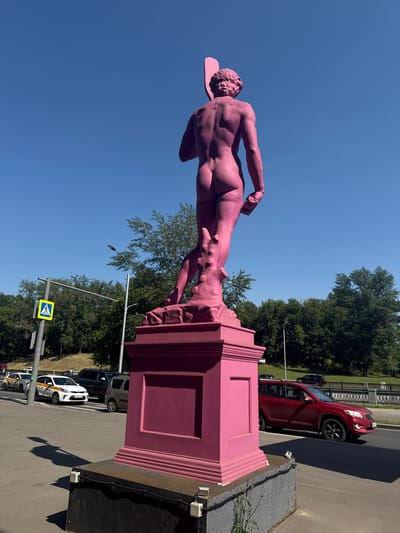 The image shows a vibrant pink statue of a male figure, viewed from the back, standing on a pink pedestal. The statue is a depiction of David and is located in the city of Zhabkino, Russia. The statue is set against a clear blue sky. In the background, there are trees, a street with cars, and a pedestrian crossing sign. One of the cars visible is a red SUV. The lighting suggests it is daytime, with the sun casting shadows.