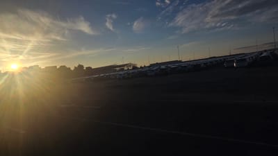 The image shows a vast outdoor car storage lot at dusk, likely a vehicle distribution center. Rows upon rows of cars, many covered with protective tarps, stretch into the distance. In the foreground, the setting sun casts a brilliant lens flare across the scene, illuminating the sky with warm hues of orange and yellow, and contrasting with the deep shadows cast by the vehicles and the earth. The sky is a mix of blue and wispy clouds, with a few tall light poles visible in the distance. The overall impression is one of a large-scale automotive inventory under a dramatic sky. The location is identified as the Pedrola, Spain.