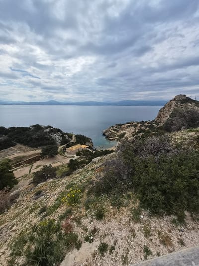 The image presents an elevated view of a coastal landscape within the Municipal Unit of Loutraki - Perachora, Greece, under an overcast sky. The foreground features a steep, arid hillside composed of light-colored rock and soil, sparsely covered with scrub brush, small green plants, and scattered yellow and white wildflowers. A grey horizontal structure is partially visible at the bottom right. In the midground, a small, tranquil bay with light blue-green water and a narrow beach is visible. A light-colored building with a reddish-brown tiled roof stands near the shoreline of the bay, accessed by a winding dirt path. To the left of the bay, an archaeological area is discernible, characterized by excavated terracing and visible stone foundations, bordered by dense green vegetation. A prominent rocky headland extends into the water on the right side of the bay. The background consists of a vast expanse of calm water, likely the sea or a gulf, extending to distant, hazy mountain ranges and landmasses beneath a sky heavily laden with grey and white clouds. No people or active subjects are present in the scene.