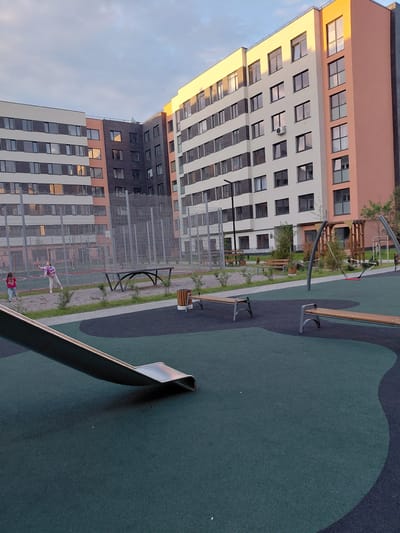 This is an outdoor photograph of a modern apartment complex playground in Guryevsk, Russia. In the foreground, a portion of a silver slide curves into a dark green rubberized surface. To the right, two dark green benches with silver metal legs are positioned on a similar surface, bordered by a lighter gray path. In the middle ground, two young girls are playing badminton on a sandy court surrounded by a chain-link fence. One girl, wearing a pink jacket and white pants, is in mid-swing. The other girl, wearing a pink shirt and blue jeans, stands nearby. Beyond the court is a long, dark gray bench with a curved design. In the background, a tall, multi-story apartment building with a beige and orange facade features numerous windows. The building is partially illuminated by the setting sun. The sky above is a mix of blue and white clouds. The scene depicts a recreational area within an urban residential environment.