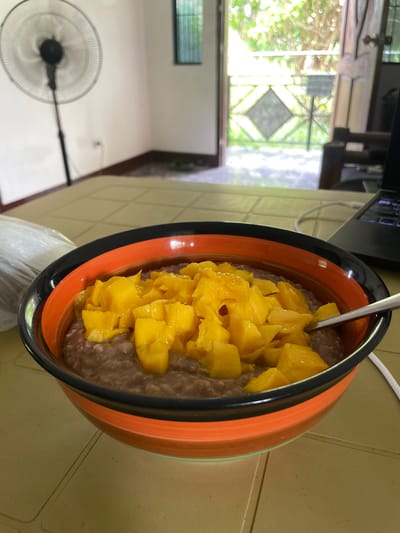 A close-up shot shows a colorful bowl of food topped with diced mangoes, placed on a light brown tiled surface. The bowl has black, orange, and green bands. A spoon rests in the bowl, partially submerged. In the background, a standing fan is visible on the left, and an open doorway leads to an outdoor area with greenery. A laptop is partially visible on the right. The scene appears to be in a residential setting, potentially in Bacong, Philippines, given the ambient lighting and foliage.