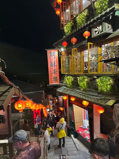 The image depicts a crowded, steeply inclined stone-paved alleyway at night in Jiufen Old Street, Ruifang District, New Taipei City, Taiwan. Numerous traditional multi-story buildings line the alley, heavily illuminated by red cylindrical and spherical lanterns strung overhead and affixed to facades, alongside decorative string lights on greenery.
On the right, a prominent building, identified by a large red banner displaying "阿妹茶樓" (A-Mei Teahouse) in black Chinese characters, features multiple floors with open yellow-framed windows and balconies adorned with luminous green foliage. Inside, several individuals are visible through the windows, seated at tables. Below this building, a storefront with a display of teaware is visible.
The alley itself is bustling with people of various ages. In the mid-ground, a person wearing a yellow overgarment carries a child on their shoulders, facing away from the camera. Another individual stands nearby, looking at a smartphone. Further down the alley, numerous figures are walking, standing, and interacting. In the lower left foreground, partially obscured, a man in a grey baseball cap and a brown top leans on a metal railing. The dark night sky dominates the upper portion of the image, revealing distant lights on the horizon, indicative of a view overlooking a bay or sea.