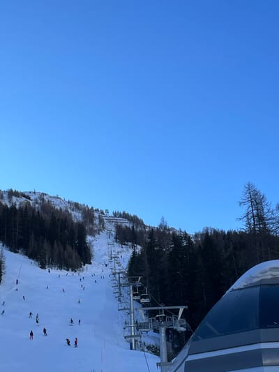 This image captures a vibrant winter scene at a ski resort, likely in Courmayeur, Italy. The sky is a clear, brilliant blue, indicating excellent weather conditions.

In the foreground, the frame is partially filled on the right by the sleek, modern design of a ski lift gondola or cabin, suggesting the photograph was taken from within or immediately adjacent to one. Extending down the slope from this point, a series of ski lift pylons and empty chairs are visible, part of the resort's transport system.

Below and to the left, a wide, groomed ski slope blanketed in white snow dominates the lower half of the scene. Numerous skiers are dispersed across the slope, engaged in various activities. Some are actively skiing down, while others appear to be standing, resting, or observing. Their colorful ski apparel adds small points of contrast against the pristine snow.

The ski slope is bordered on its left by a mountainside densely covered with trees. A mix of dark evergreen trees and some bare deciduous trees are visible, extending up the incline. The overall impression is one of a busy and well-maintained ski area under clear skies, likely during the daytime. No visible text is discernible in the image.