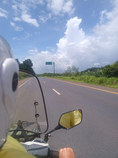 The image, likely a point-of-view shot, shows a person riding a motorcycle on a paved road. The rider is wearing a white helmet and a yellow shirt or jacket. The road has a yellow line on the right edge and broken white lines dividing lanes. A signpost is visible on the left side of the road with writing in Thai and English, indicating "U-TURN 1 KM". The environment consists of green vegetation and trees alongside the road. The sky is blue with scattered white and gray clouds. The reflection in the motorcycle's side mirror reveals a crumpled yellow material, possibly a rain jacket or plastic bag. Given the signpost stating "U-TURN 1 KM," it's plausible that the scene is located near Plai Phraya, Thailand.