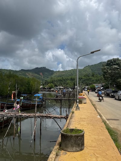 In Ban Hin Rom, Thailand, a cloudy day casts shadows over a waterfront scene. A narrow waterway runs alongside a road, with wooden piers and traditional longtail boats moored on the left. The water reflects the overcast sky and the surrounding mangrove trees. On the right, a paved yellow walkway runs parallel to the road, bordered by sparse grass. A man on a motorcycle rides away from the camera on the road, and parked cars are visible further down. In the background, lush green hills rise, with a noticeable landslide scar on one slope. A street lamp stands prominently on the walkway.