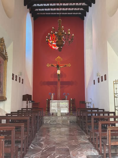 This media file captures the interior of a quiet church or chapel in San Lucas el Grande, Mexico. The scene is devoid of people or activity, conveying a sense of stillness and reverence.
The central focal point is a large, ornate golden crucifix depicting Jesus, prominently mounted against a vibrant red accent wall. Above Jesus' head, the inscription "INRI" is clearly visible. To the left of the crucifix hangs a grand, multi-tiered chandelier with numerous bulbs and a circular stained-glass section that casts warm, colorful light.
The architecture of the space includes clean white walls and a ceiling with graceful, curved sections and several recessed light fixtures. Along the viewer's right side, multiple rows of dark wooden pews are neatly arranged, facing the altar. The floor and the wall directly behind the pews are covered in distinctive marbled tiles, predominantly in tones of reddish-brown with white veining.
In the altar area, a simple, light-colored rectangular altar or table is present, with a small vase holding purple flowers positioned behind it. Other visible details include dark wooden beams on the left side of the room, an arched window on the left wall providing subtle natural light, and the top portion of a large, gold-framed painting in the lower right foreground. Small rectangular speakers or vents are mounted on the white ceiling/wall. The overall ambiance suggests a place of traditional religious worship and quiet contemplation.