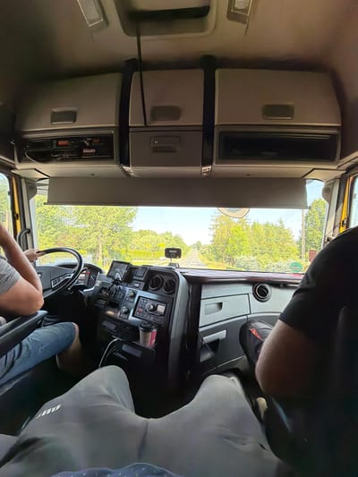 This media file captures an interior view from the driver's seat of a truck, looking out through the windshield and side window. The driver, whose legs and arms are visible, is seated at the steering wheel, suggesting they are either driving or about to drive. To the left of the driver, there are several cabinets and storage compartments built into the truck's cabin, typical of a long-haul or commercial vehicle. The dashboard features a complex array of buttons, switches, and displays, including a GPS or navigation screen. A red cup is visible in a holder on the dashboard.

Through the windshield and side window, a sunny, tree-lined road is visible, indicating an outdoor daytime scene. The greenery suggests a temperate climate and possibly a rural or suburban setting. The sky appears bright, suggesting pleasant weather.

The scene appears to be set in Mordarka, Poland, as per the provided location context. There are no overt emotional cues from the visible subjects, and the activity is primarily the operation of a vehicle. The focus is on the truck's interior and the view outside. A green circular sign with a white number is visible on a pole outside the truck, but the number is not clearly legible. A smaller black rectangular sign is also visible on the dashboard.