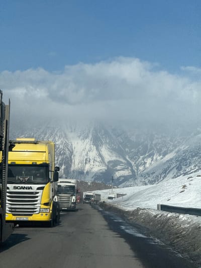 An outdoor, daytime image captures a line of heavy-duty trucks on an asphalt road in a snow-covered mountainous region, located in the city of Kanobi, Georgia. In the immediate foreground on the left, the front of a yellow Scania R480 semi-truck is visible, featuring a prominent grille and displaying a Georgian license plate "522 LL 22" on its bumper. The road surface exhibits wet patches, bordered by significant snowbanks on either side. A metal guardrail runs along the right edge of the road in the midground. Extending into the distance, a queue of multiple semi-trucks, including white Scania and Ssangyong models, are stationary or moving slowly on the road. The background is dominated by large, rugged mountains, heavily covered in snow and ice, with their peaks partially obscured by low-lying white clouds. A clear blue sky is visible above the cloud line. Small structures or shacks are discernible along the right side of the road in the midground. No specific individuals are clearly identifiable outside the vehicles.
