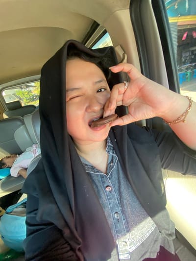 A woman wearing a black hijab and a denim shirt is sitting in the passenger seat of a car and eating a chocolate biscuit. She is winking at the camera. A child is asleep in the back seat of the car. The car appears to be stationary, possibly parked on the side of a road in Jotangan, Indonesia. The environment outside the car suggests a commercial or residential area with buildings and trees visible.