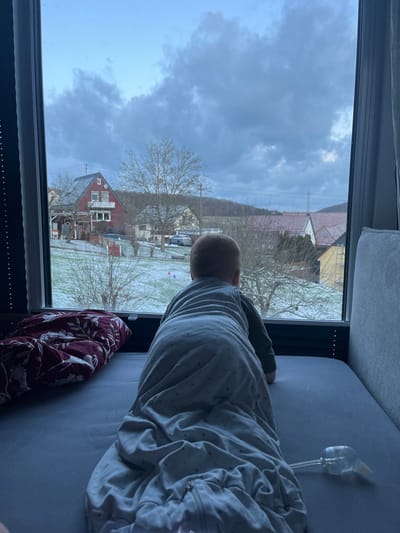 A child in a sleep sack looks out a window at a snowy landscape. Outside, a red house with a white facade and bare trees are visible. The sky is overcast with heavy, dark clouds. A bottle with a tube attachment rests on the bed next to the child. A patterned blanket is draped on the bed. The photo is taken from a slightly elevated position.