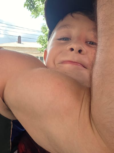 The media file shows a close-up, upside-down, low-angle selfie of a young boy, likely a child or pre-teen. He has fair skin, light-colored eyes, and dark eyebrows. He is wearing a dark baseball cap on his head, with the brim partially obscuring his forehead. The boy appears to be outdoors, as indicated by the bright sky and greenery visible in the background, suggesting it is daytime. The overall mood seems playful and casual, given the selfie style and the boy's slightly amused expression. The background hints at a residential area with trees and what looks like part of a building or fence. There is no visible text in the image. The location context provided is Krymsk, Russia, which helps contextualize the outdoor setting.