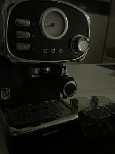 A dark, close-up shot features a black and silver espresso machine. The left side showcases a pressure gauge and three buttons. A chrome-plated knob sits atop the machine. Adjacent to it is a portafilter and on the right is a drip tray with many small holes. The word "GASTROBACK" is visible on the far right edge of the machine. The lower corner has a QR code and text that begins "VIDEO &...". The lighting is dim, highlighting the machine's metallic accents.