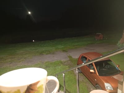 A nighttime outdoor scene is depicted with a bright full moon visible in the dark sky above a body of water and silhouetted trees. In the foreground, a white mug with a floral design sits on the left, partially obscuring a metal railing and stairs. To the right of the railing, an orange hatchback car is parked on a grassy area. Further in the background, a small, reddish-brown wooden structure, possibly a doghouse or playhouse, is situated on the grass. The environment appears to be rural, consistent with a location in Maļutki, Latvia.