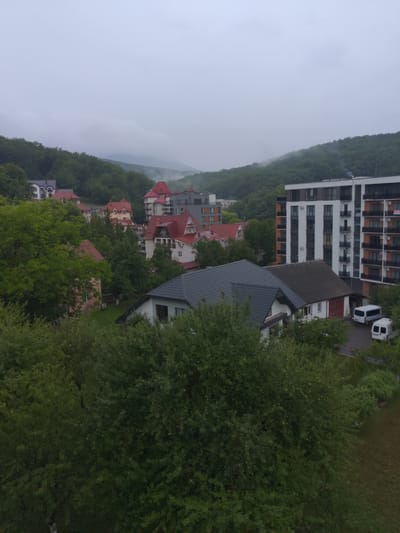 The image shows a view of a residential area nestled among lush green trees, with hills in the background under an overcast sky. The architecture visible includes several multi-story buildings with balconies and red-tiled roofs, typical of European residential development. Some smaller houses with red roofs are also present. Two white vans are parked on an asphalt surface near the buildings, suggesting a community or a resort area. The overall scene depicts a tranquil, possibly damp, day in a town or village, with nature prominently featured alongside human structures. The location is identified as Сонячне, Ukraine.