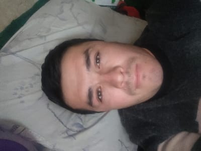 An adult male is depicted in a close-up, self-portrait style image, captured indoors in the city of Yangikurgan, Uzbekistan. The individual, appearing to be of Central Asian descent, possesses dark hair and brown eyes. He exhibits stubble on his chin and upper lip. He is wearing a dark, long-sleeved garment, likely a sweater or shirt, which covers his upper torso. His head rests on a light-colored pillow featuring a subtle, repeating patterned design, possibly floral or abstract, in grey or blue tones. A dark green fabric edge is partially visible in the upper left background, suggesting the presence of additional bedding or furnishing. The male is positioned horizontally, looking directly at the camera with a neutral expression. No other individuals or interactions are visible. The environment appears to be an interior space, likely a bedroom or living area.