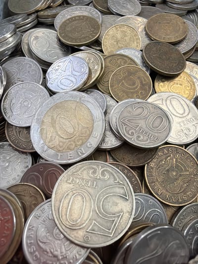 The image is a close-up, high-angle, overhead shot of a large pile of assorted coins. The coins are mostly round and appear to be made of metal, with various denominations and designs visible. Many of the coins are silver or a brassy-gold color, and some show signs of wear and age. The coins are stacked and jumbled together, creating a textured surface with many overlapping edges. Text and numerals are visible on some of the coins, suggesting they are currency. The environment is not discernible beyond the coins themselves, but the coins themselves are from Kazakhstan, and thus are likely found in the city of Şalqar, Kazakhstan.
