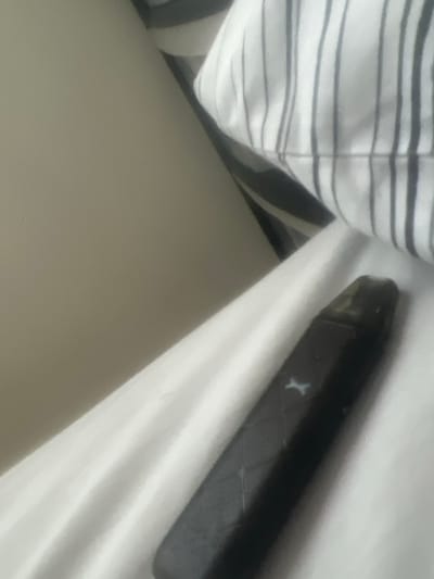 A black, elongated vaping device with a textured surface and a light-colored 'X' emblem near its top rests on white, rumpled bed linen. The device features a translucent dark mouthpiece. To the upper right, a pillow or folded fabric with vertical black and white stripes is partially visible. The upper left background consists of a plain, light beige wall or headboard. No individuals are present, and no actions or interactions are depicted. The scene represents an indoor environment, likely a bedroom. The image contains no identifiable features to confirm a location within Southampton, United Kingdom.