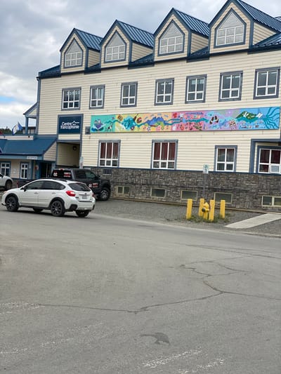 The image captures the exterior of the Centre de la Francophonie in Whitehorse, Canada, on an overcast day. The building features a light-colored, horizontally-paneled facade with dark blue trim around the gabled roof sections and windows. A large, colorful mural depicting various scenes, including natural landscapes and stylized figures, runs across the mid-section of the building.
In front of the building, a white Subaru Outback is parked and facing to the left. A dark-colored pickup truck, possibly a Ford F-series, is parked behind the Subaru and to the right, with "CODY'S" branding visible on its door. The ground in front of the building is a mix of asphalt and gravel. A yellow fire hydrant with protective bollards stands near the right side of the frame, accompanied by a "P" parking sign. The sky is cloudy, suggesting a temperate climate. The general environment appears to be an urban or semi-urban setting, likely in a northern location given the building's design and the context of Whitehorse being a Canadian city.