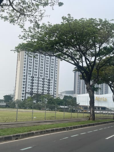 The image depicts a street view in Pakulonan Barat, Indonesia, likely in the daytime under overcast skies. In the background, a tall, modern apartment building with a white and grey facade, featuring numerous rectangular windows, dominates the skyline. To its right, another similar building is visible, and further in the distance, parts of industrial structures and a crane can be observed. In the foreground, a large tree with dense green foliage stands prominently on the right side of the road. A metal fence separates a grassy field from the road. The road itself is paved and shows lane markings. A white building with the word "Atria" in golden lettering is situated behind the fence. There are no people visible in the image.