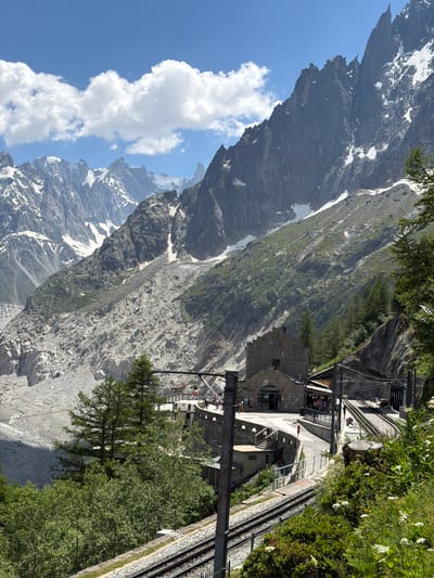 This image depicts a dramatic mountain landscape with a train station nestled on a steep incline. The primary subjects are the rugged, snow-dusted mountains that dominate the background, a stark contrast to the lush green trees and vegetation in the foreground. A stone building, likely part of the train station, is visible with people gathered on its platforms and walkways. Train tracks snake along the side of the mountain, leading to and from the station. The scene appears to be set on a sunny day, with blue skies and white clouds visible. The steepness of the terrain suggests a mountainous region, and the presence of snow on the peaks indicates a high-altitude location. The overall impression is one of a majestic natural environment integrated with human infrastructure, likely a tourist destination. No visible text is present in the image.