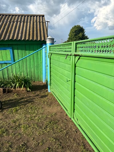 A bright green metal gate with a star-shaped pattern on its upper section stands to the right of the image. The gate is partially open, revealing the interior of a yard. To the left of the gate, a green wooden building with a corrugated roof is visible. A blue window frame is present on the building's wall. In front of the building, a wooden planter box holds lush green plants. The ground is a mix of dirt and sparse grass, with shadows cast by the objects in the scene. The sky above is partly cloudy with dramatic clouds. The scene appears to be in an outdoor rural or semi-rural setting. The location is identified as Чуй-Атасево, Russia.
